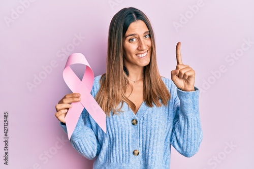 Brunette young woman holding pink cancer ribbon smiling with an idea or question pointing finger with happy face, number one