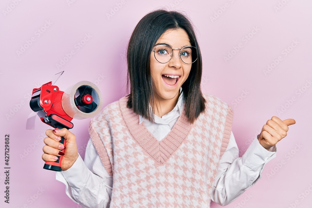 Young brunette woman holding packing tape pointing thumb up to the side smiling happy with open mouth Young brunette woman holding packing tape pointing thumb up to the side smiling happy with open mouth
