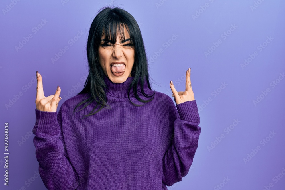 Young brunette woman with bangs wearing turtleneck sweater shouting with crazy expression doing rock symbol with hands up. music star. heavy concept. Young brunette woman with bangs wearing turtleneck sweater shouting with crazy expression doing rock symbol with hands up. music star. heavy concept.