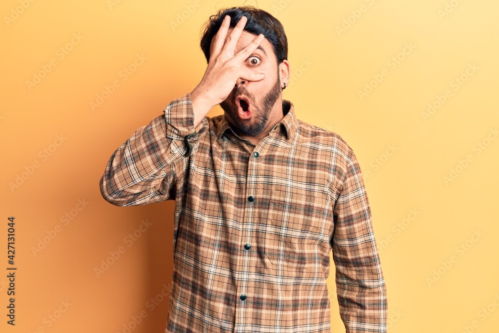 Young hispanic man wearing casual clothes peeking in shock covering face and eyes with hand, looking through fingers afraid Young hispanic man wearing casual clothes peeking in shock covering face and eyes with hand, looking through fingers afraid