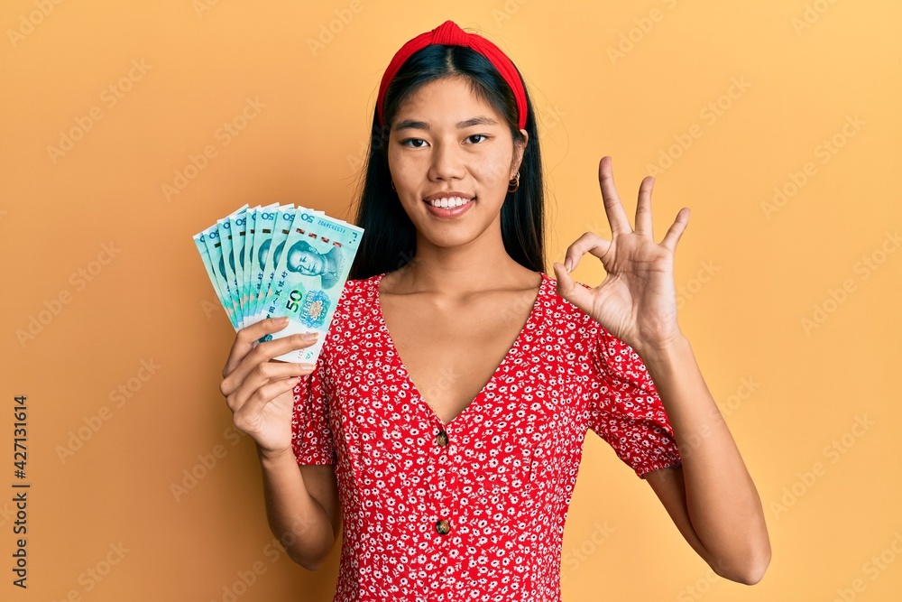 Young chinese woman holding 50 yuan chinese banknotes doing ok sign with fingers, smiling friendly gesturing excellent symbol