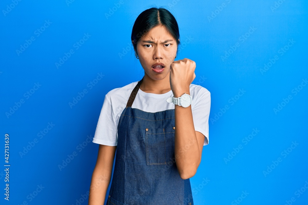 Young chinese woman wearing waiter apron angry and mad raising fist frustrated and furious while shouting with anger. rage and aggressive concept. Young chinese woman wearing waiter apron angry and mad raising fist frustrated and furious while shouting with anger. rage and aggressive concept.