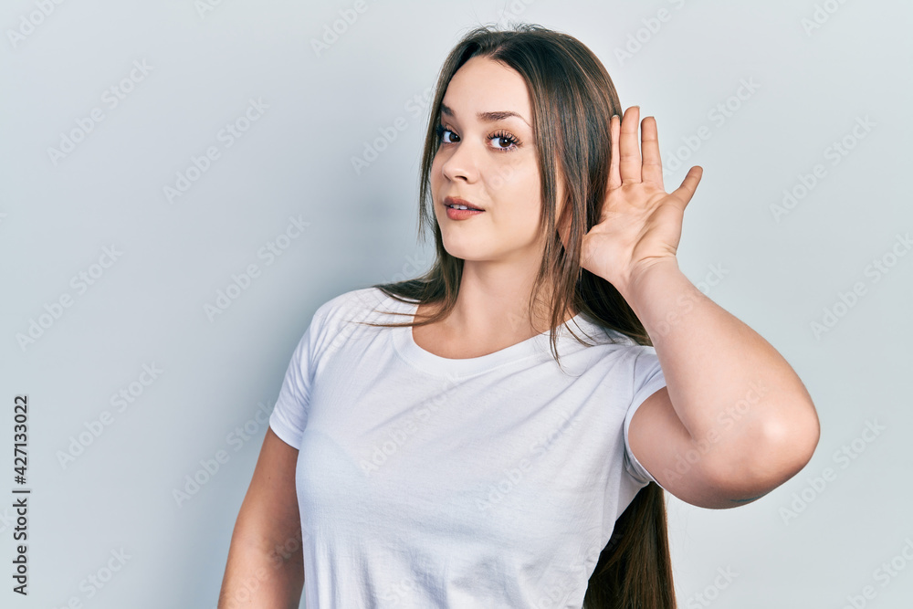 Young hispanic girl wearing casual white t shirt smiling with hand over ear listening and hearing to rumor or gossip. deafness concept. Young hispanic girl wearing casual white t shirt smiling with hand over ear listening and hearing to rumor or gossip. deafness concept.