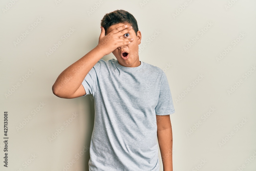 Teenager hispanic boy wearing casual grey t shirt peeking in shock covering face and eyes with hand, looking through fingers with embarrassed expression.