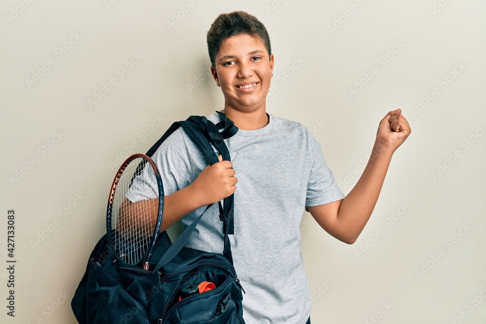 Teenager hispanic boy holding sport bag screaming proud, celebrating victory and success very excited with raised arm