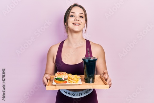 Young hispanic girl wearing sportswear eating a tasty classic burger with fries and soda smiling and laughing hard out loud because funny crazy joke.