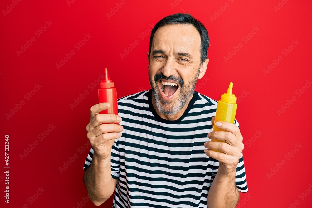 Middle age hispanic man holding ketchup and mustard bottle smiling and laughing hard out loud because funny crazy joke. Middle age hispanic man holding ketchup and mustard bottle smiling and laughing hard out loud because funny crazy joke.