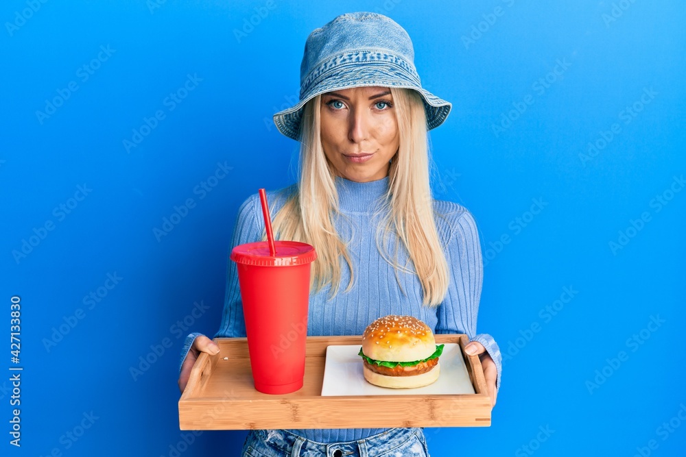 Young blonde woman eating a tasty classic burger and soda relaxed with serious expression on face. simple and natural looking at the camera. Young blonde woman eating a tasty classic burger and soda relaxed with serious expression on face. simple and natural looking at the camera.