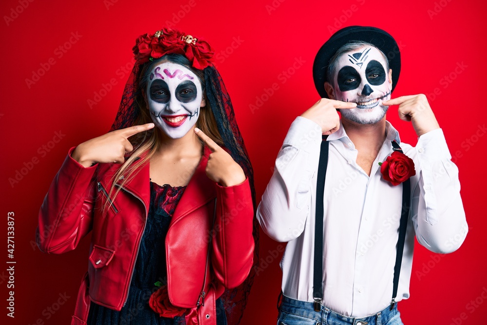 Couple wearing day of the dead costume over red smiling cheerful showing and pointing with fingers teeth and mouth. dental health concept. Couple wearing day of the dead costume over red smiling cheerful showing and pointing with fingers teeth and mouth. dental health concept.