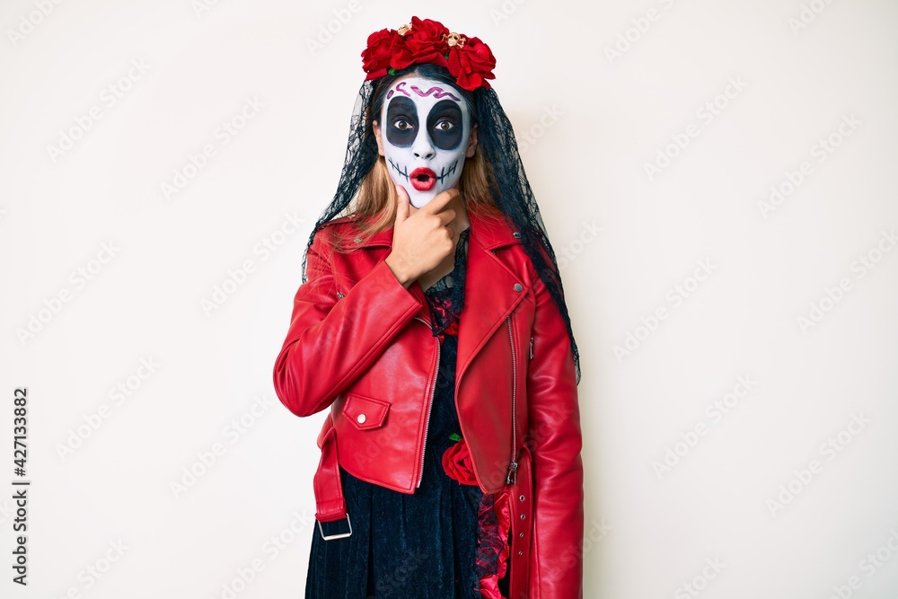 Woman wearing day of the dead costume over white looking fascinated with disbelief, surprise and amazed expression with hands on chin Woman wearing day of the dead costume over white looking fascinated with disbelief, surprise and amazed expression with hands on chin
