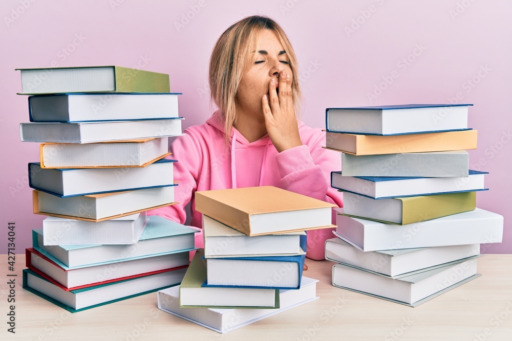 Young caucasian woman sitting on the table with books bored yawning tired covering mouth with hand. restless and sleepiness. Young caucasian woman sitting on the table with books bored yawning tired covering mouth with hand. restless and sleepiness.