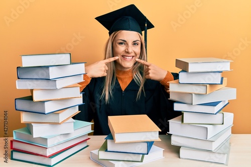 Young caucasian woman wearing graduation ceremony robe sitting on the table smiling cheerful showing and pointing with fingers teeth and mouth. dental health concept.