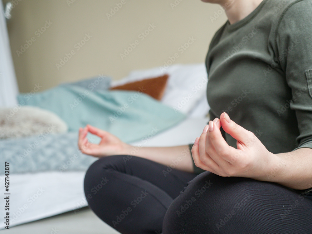 Fototapeta premium Woman sitting on floor in posture of meditation close up. Girl doing meditative practice in lotus position in bedroom. Active life in self-isolation.