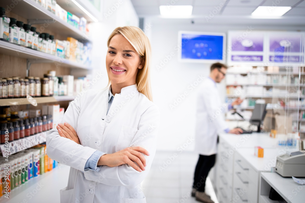 Portrait of beautiful blonde female pharmacist standing in pharmacy store by the shelf with medicines.