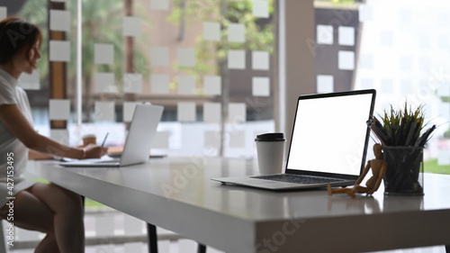 Mock up computer laptop with blank screen white table and businesswoman sitting in background. © Prathankarnpap