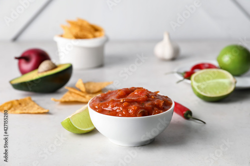 Bowl of tasty salsa sauce with ingredients and nachos on light background