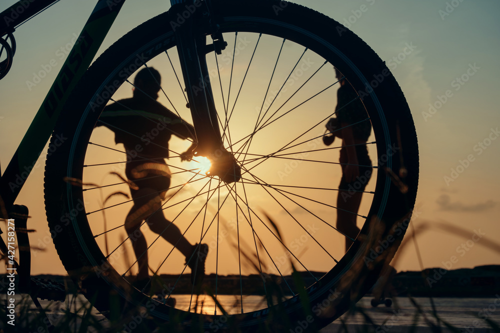 Close-up silhouette of a bike wheel at sunset. The sun shines through the wheel of a bicycle with blurred  runner figure at background