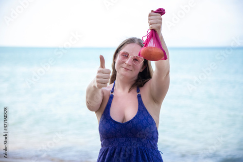 smiling caucasian woman holding mesh bag with fruit and showing thumbs up. on the background is sea or ocean.