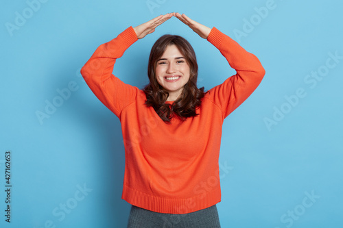 Smiling dark haired girl wearing casual style outfit making roof from her head and looking at camera with pleasant smile, wants to be in safety, isolated over blue background. © sementsova321