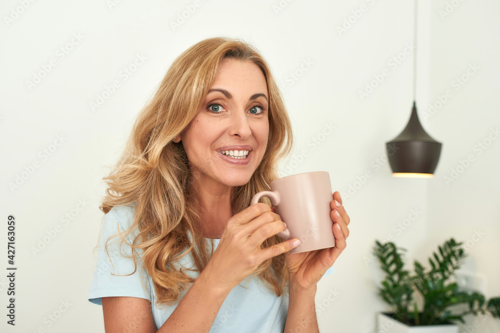 A portrait of a beautiful middle-aged smiling into the camera woman on a self-isolation with a cup. A portrait of a beautiful middle-aged smiling into the camera woman on a self-isolation with a cup.