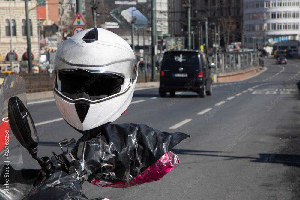 In the focus in the foreground is a white, worn female motorcycle helmet attached to the handlebars of a motorbike. The photo was taken on a wide asphalt street in a big city. In the focus in the foreground is a white, worn female motorcycle helmet attached to the handlebars of a motorbike. The photo was taken on a wide asphalt street in a big city.