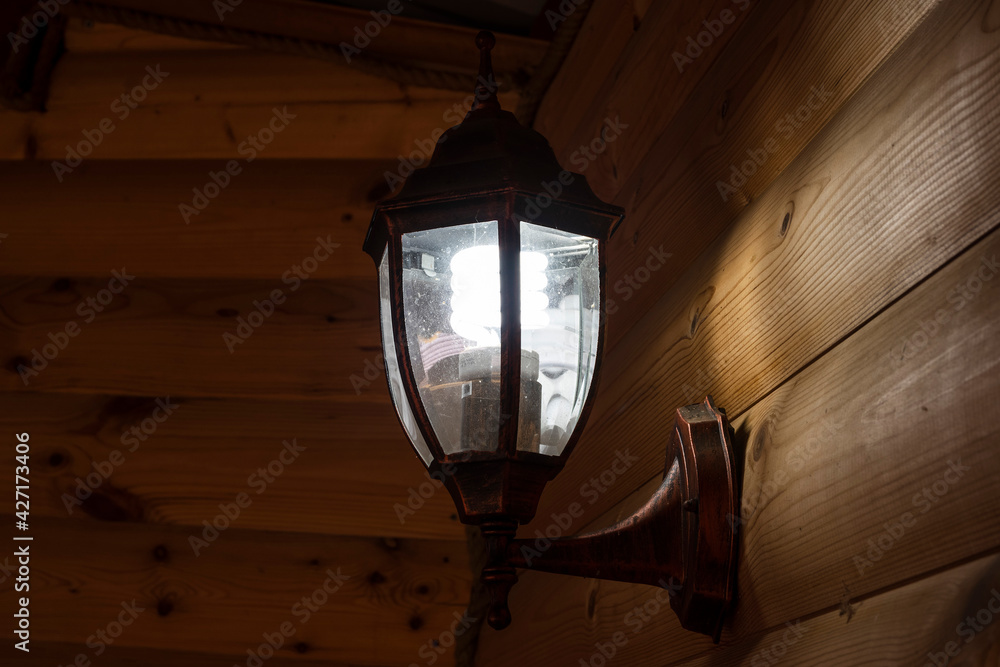 A metal brown street lamp with an energy-saving lamp on the log wall of the house under the canopy shines in the evening. A way to create a romantic setting on the porch of your home.