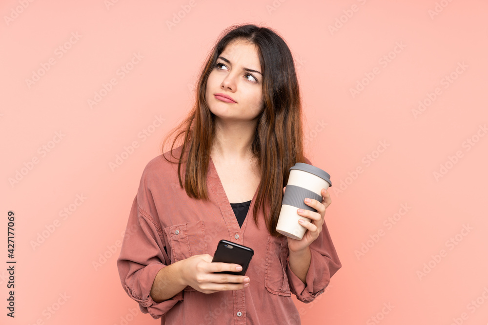Young caucasian woman isolated on pink background holding coffee to take away and a mobile while thinking something Young caucasian woman isolated on pink background holding coffee to take away and a mobile while thinking something
