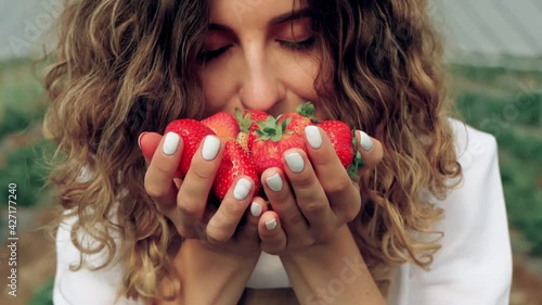 Closeup crop motion of smiling with teeth woman with curly hair carrying strawberries in hand palms. Young caucasian female farmer wearing apron smelling scent of ripe organic berries at greenhouse.