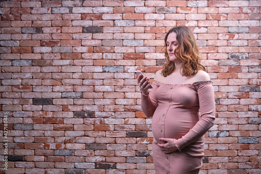 Portrait of a pregnant woman with a mobile phone in her hands against a background of a brick wall. An adult woman looking at the phone holding her belly