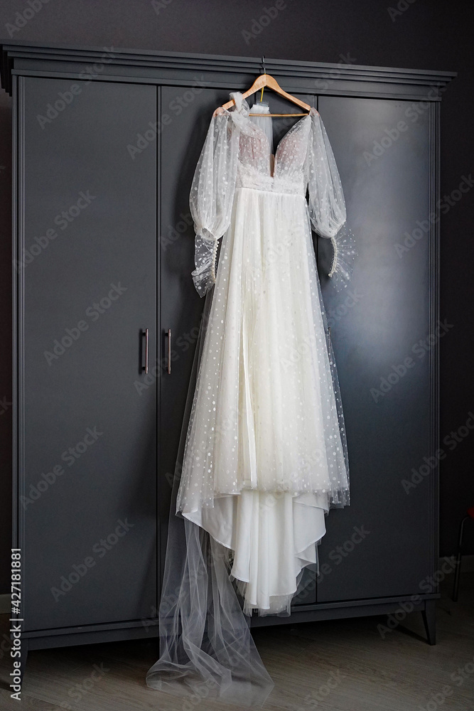 A long white beautiful wedding dress hangs on the door of a dark closet. A long white beautiful wedding dress hangs on the door of a dark closet.