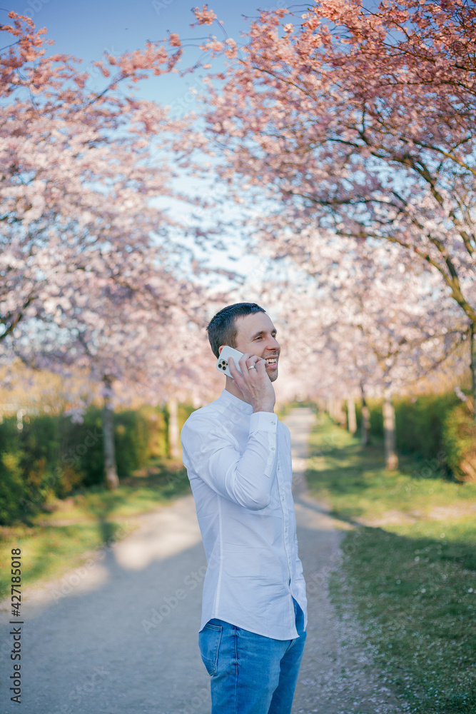 A young man speaks on the phone against the background of a blossoming sakura garden. Spring. A young man speaks on the phone against the background of a blossoming sakura garden. Spring.