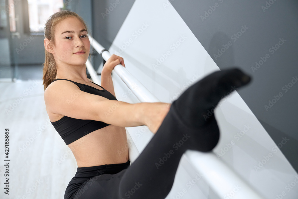 Close up of attractive teenager holding leg on handrail. Flexible female athlete wearing tight black sports clothes practicing split in empty dance hall with hi tech interior. Concept of gymnastics. Close up of attractive teenager holding leg on handrail. Flexible female athlete wearing tight black sports clothes practicing split in empty dance hall with hi tech interior. Concept of gymnastics.