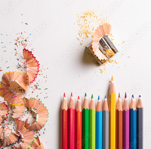 Top view of the colored pencils side by side highlighting the yellow pencil on the white background. With colored pencil shavings on the left and a pencil sharpener on the upper right.