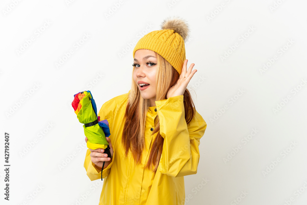 Teenager blonde girl wearing a rainproof coat over isolated white background listening to something by putting hand on the ear Teenager blonde girl wearing a rainproof coat over isolated white background listening to something by putting hand on the ear