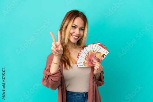Teenager blonde girl taking a lot of Euros over isolated blue background smiling and showing victory sign