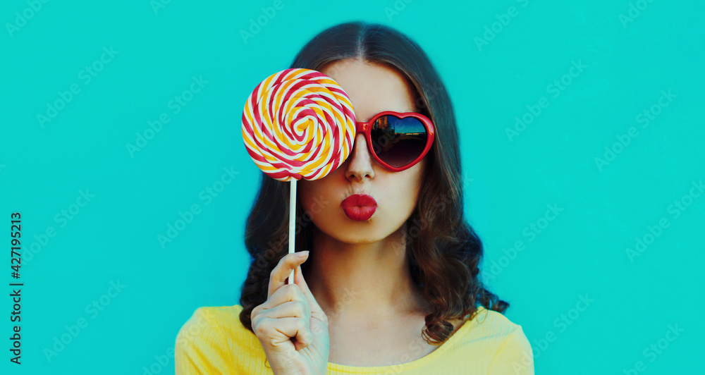 Portrait close up of young woman blowing her red lips with lollipop on a blue background