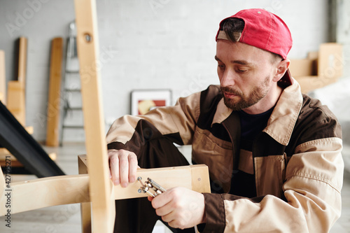 Принти на полотні Serious young furniture assemblage specialist in workwear fixing parts of chair