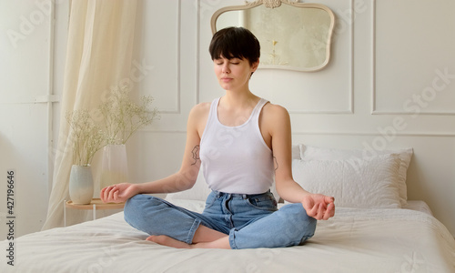Young woman meditating while sitting on the bed at home in bedroom. Calm female taking deep breath and relaxing with eyes closed. No stress and peace of mind.