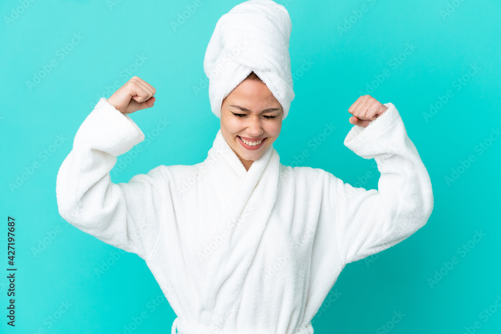 Teenager blonde girl in a bathrobe over isolated blue background doing strong gesture Teenager blonde girl in a bathrobe over isolated blue background doing strong gesture