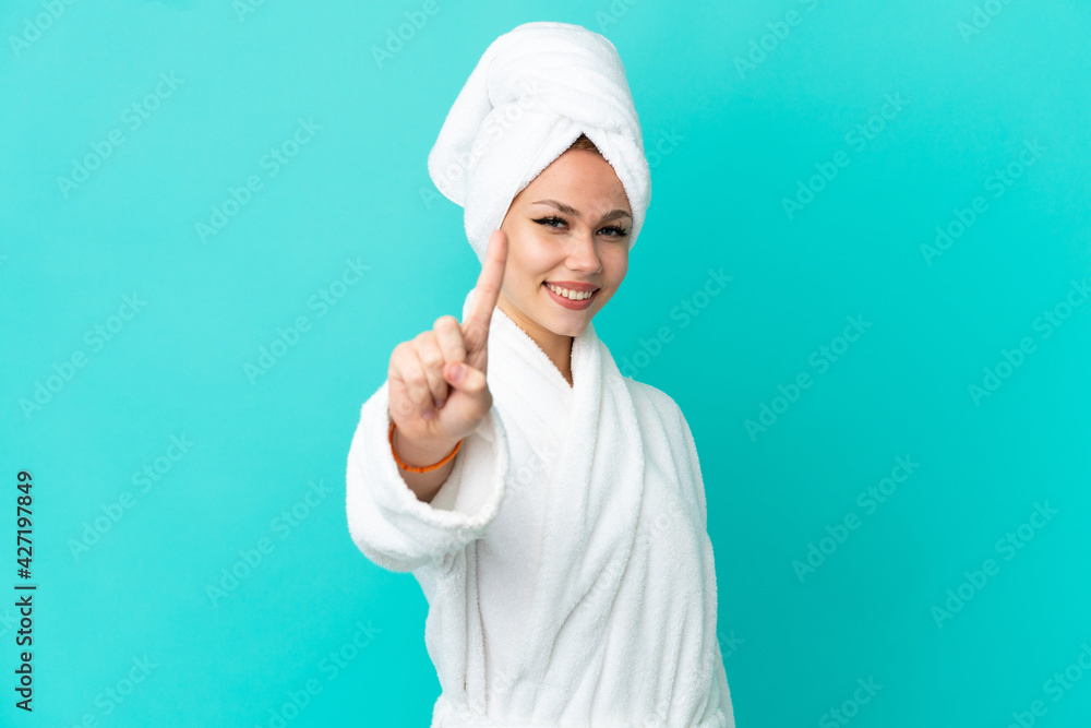 Teenager blonde girl in a bathrobe over isolated blue background showing and lifting a finger Teenager blonde girl in a bathrobe over isolated blue background showing and lifting a finger