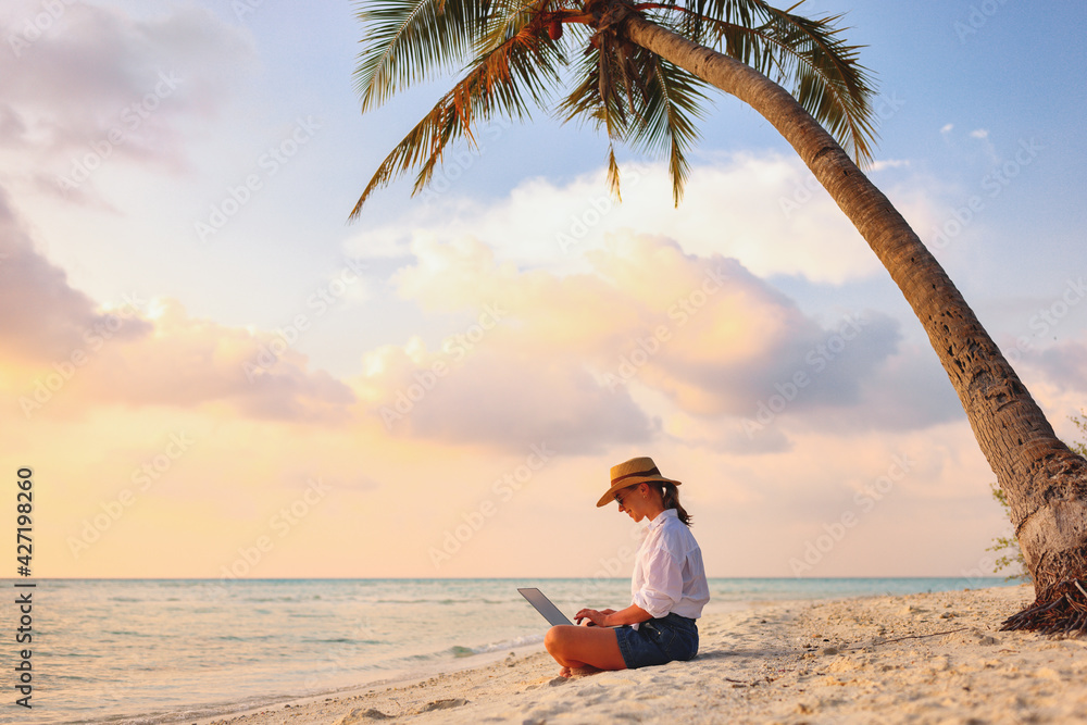 Young female freelancer wearing straw hat working on laptop while sitting on tropical beach at sunset