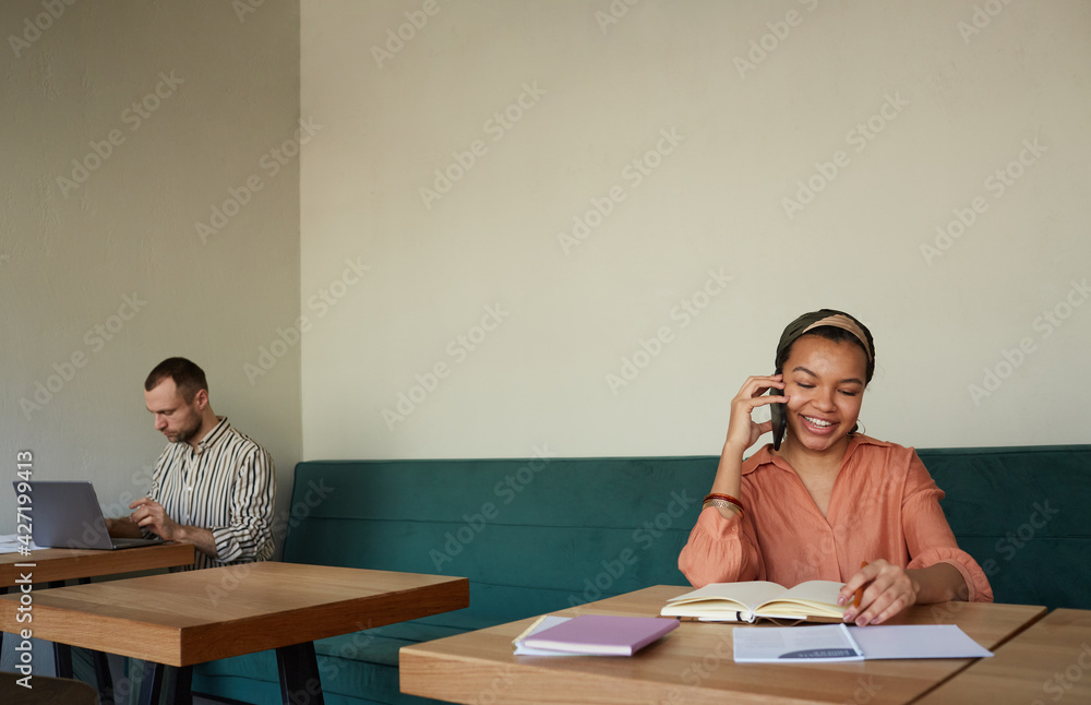 Minimal shot of two people working at tables in cafe focus on smiling African-American businesswoman speaking by smartphone, copy space Minimal shot of two people working at tables in cafe focus on smiling African-American businesswoman speaking by smartphone, copy space