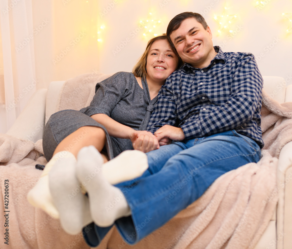 portrait of adult romantic couple sitting on a couch at home, holiday lights on a wall portrait of adult romantic couple sitting on a couch at home, holiday lights on a wall