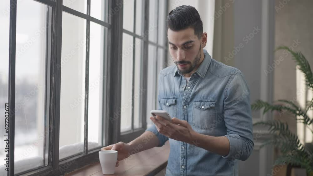 Confident handsome man drinking morning coffee and messaging online on smartphone. Portrait of Middle Eastern guy surfing social media at home indoors. Modern lifestyle Confident handsome man drinking morning coffee and messaging online on smartphone. Portrait of Middle Eastern guy surfing social media at home indoors. Modern lifestyle