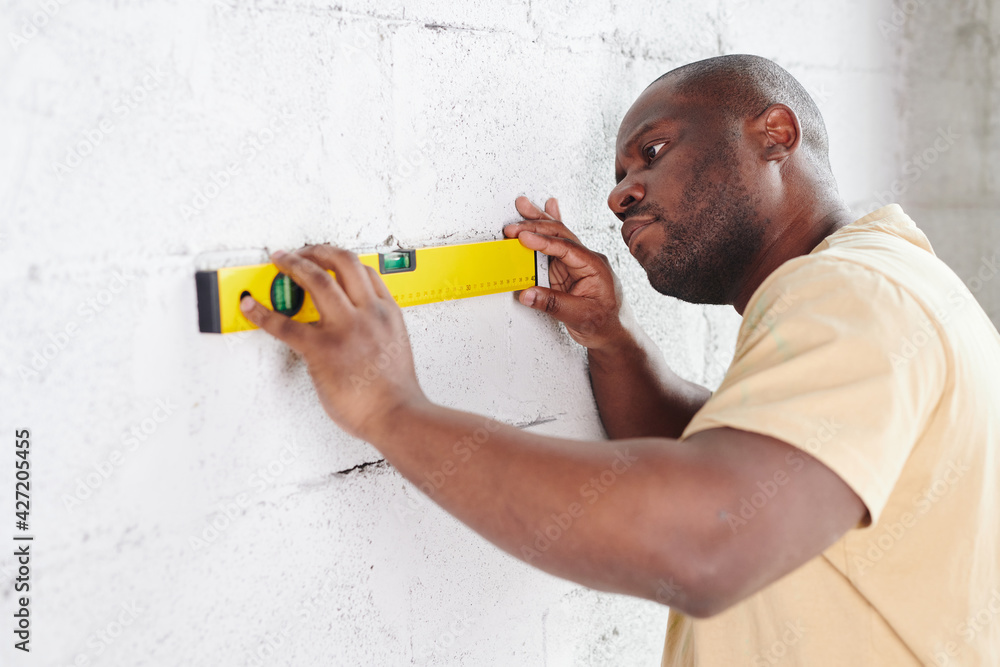 Young man of African ethnicity putting level measuring hand tool to white painted brick wall while checking its flatness during home renovation Young man of African ethnicity putting level measuring hand tool to white painted brick wall while checking its flatness during home renovation