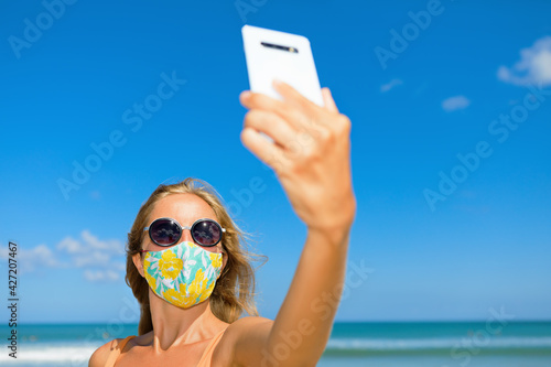 Funny girl taking selfie photo by smartphone on tropical sea beach. New rules to wear cloth face covering mask at public places due coronavirus COVID 19. Family holidays with children, summer travel. © Tropical studio