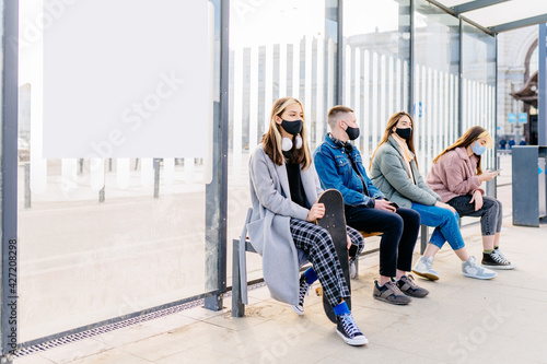 Group of young male and females people im protective masks sitting in line at bus stop. Keeping safe social distance. Hipsterh man and women outdoor waiting for transport.