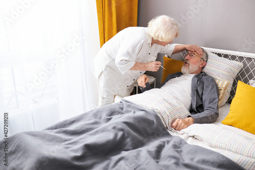 Woman take care of sick husband, checking fever temperature of senior man lying on bed at home photo