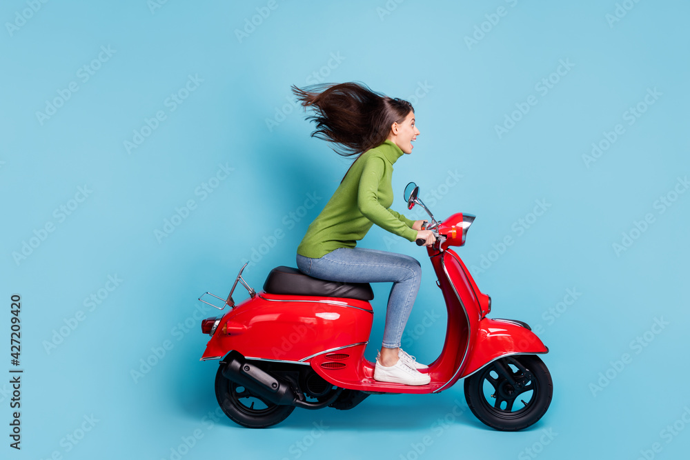 Full length side photo photo portrait of excited girl driving red scooter isolated on pastel blue colored background Full length side photo photo portrait of excited girl driving red scooter isolated on pastel blue colored background