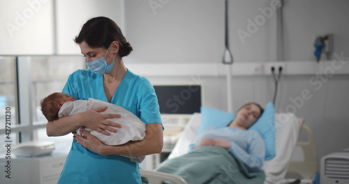 Nurse in medical mask holding newborn baby with mother resting in bed on background